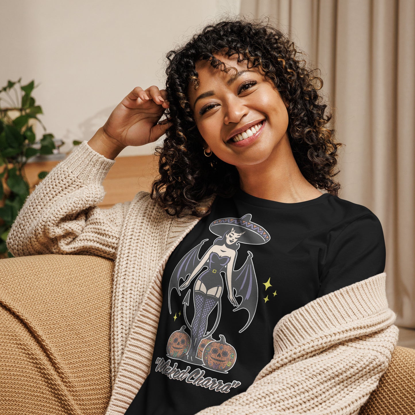 Smiling woman wearing a 'Wicked Charra' black t-shirt with a retro pinup charra design, including devil wings, a sombrero, and two jack-o-lanterns. The t-shirt combines Halloween and Día de los Muertos themes.