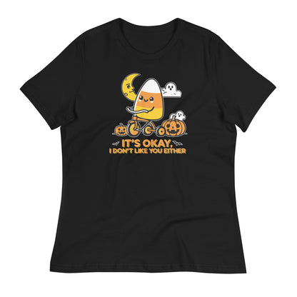 A flat-lay of a black t-shirt featuring the same design of a candy corn character on a tricycle with pumpkins, ghosts, and the phrase "It's okay, I don't like you either.