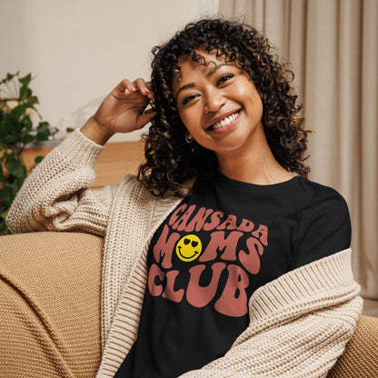 A young woman sitting on a coach wearing a knitted sweater with a black tshirt with the words Cansada Moms Club with a smiley face and hearts.